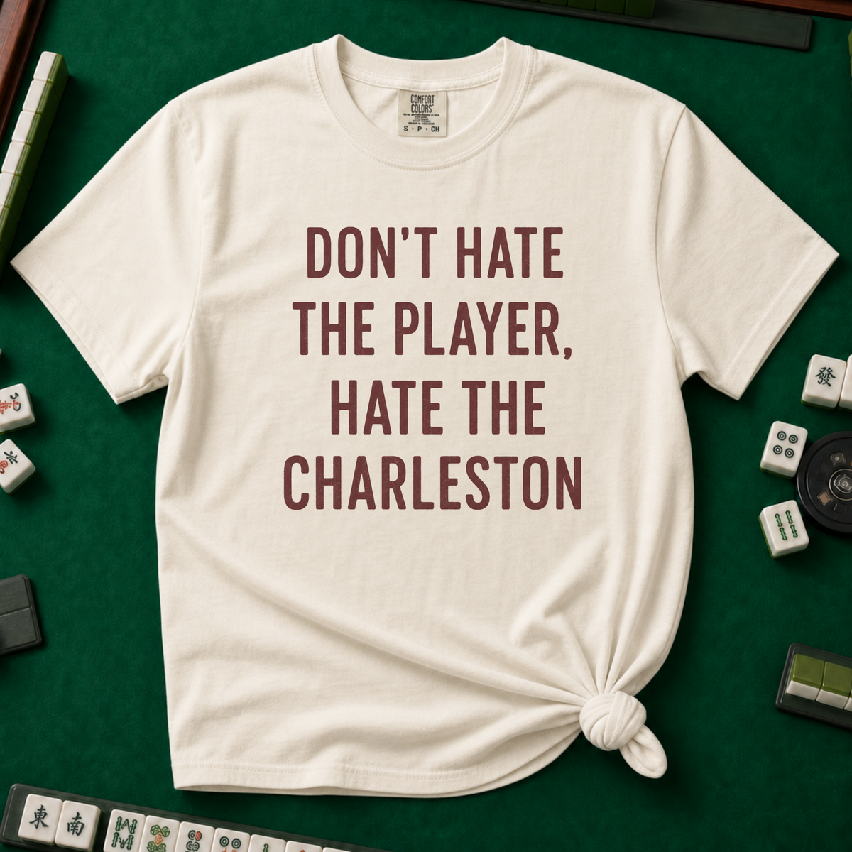 Don't Hate The Player Hate The Charleston T-Shirt in Ivory, knotted flat lay on a green mahjong mat - "DON'T HATE THE PLAYER, HATE THE CHARLESTON" in deep burgundy block letters
