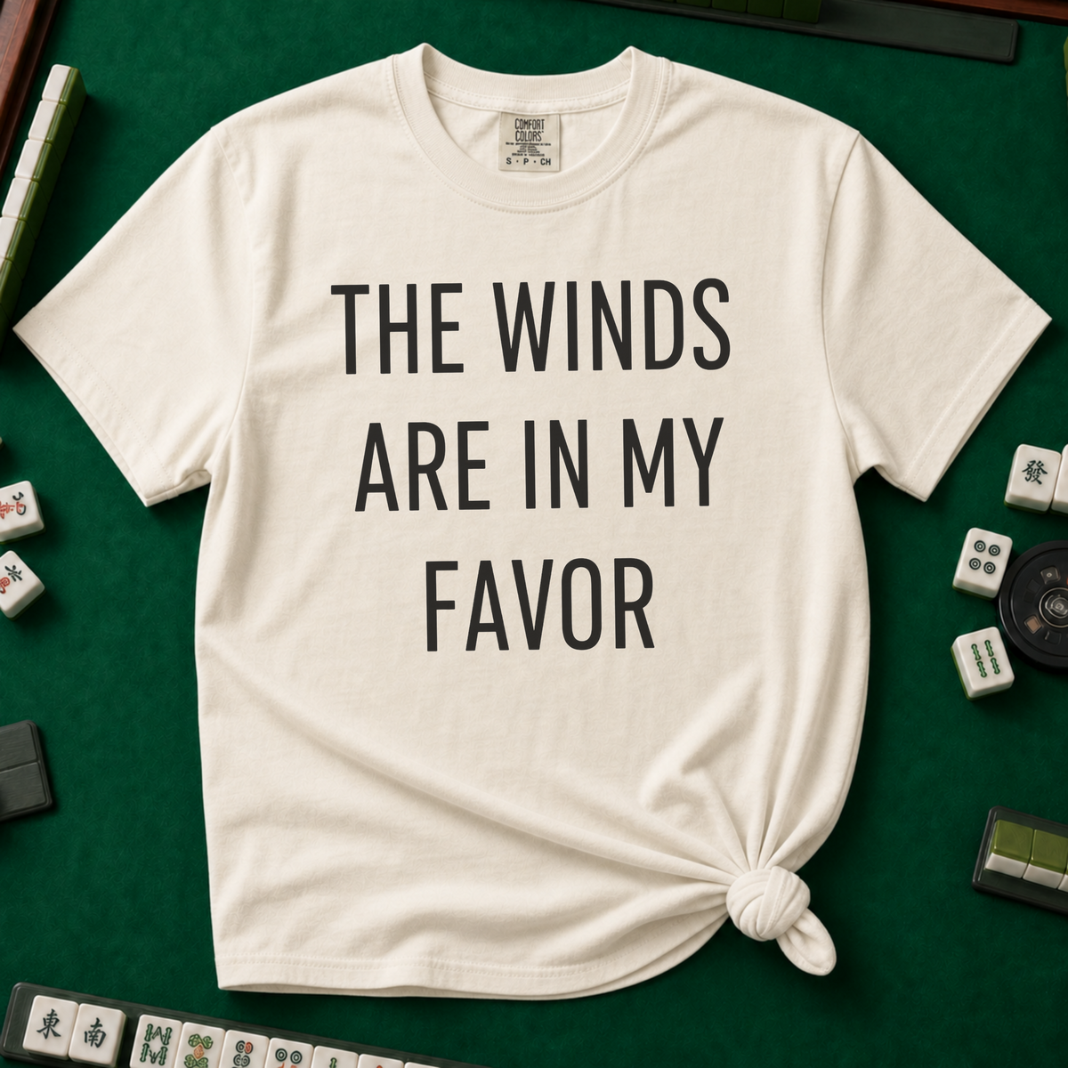 The Winds Are In My Favor T-Shirt in Ivory, knotted flat lay on a green mahjong mat - "THE WINDS ARE IN MY FAVOR" in tall black letters