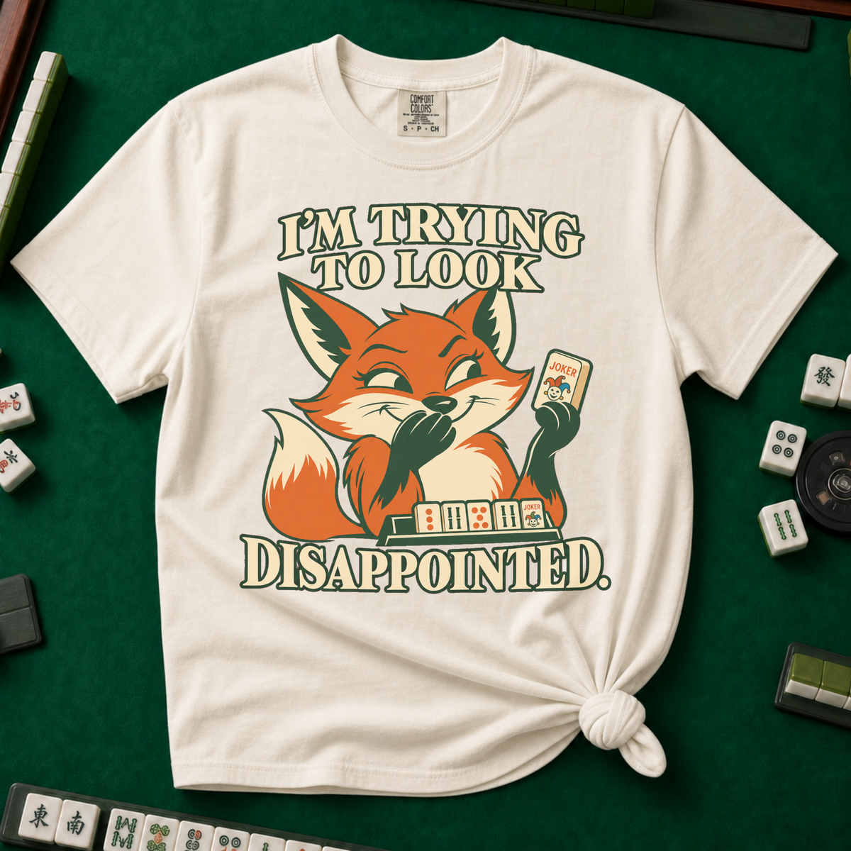 I'm Trying To Look Disappointed T-Shirt in Ivory, knotted flat lay on a green mahjong mat - smug retro fox holding up a joker tile, hiding a grin behind one paw, tile rack in front - "I'M TRYING TO LOOK DISAPPOINTED." in cream collegiate letters