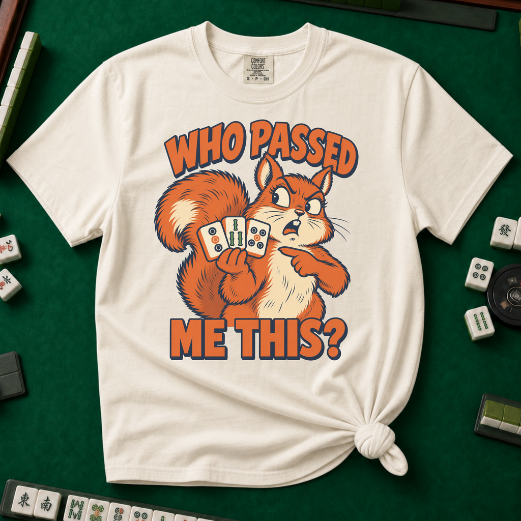 Who Passed Me This T-Shirt in Ivory, knotted flat lay on a green mahjong mat - retro orange squirrel holding three mahjong tiles, pointing an accusing paw - "WHO PASSED ME THIS?" in bold orange letters