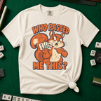 Who Passed Me This T-Shirt in Ivory, knotted flat lay on a green mahjong mat - retro orange squirrel holding three mahjong tiles, pointing an accusing paw - "WHO PASSED ME THIS?" in bold orange letters