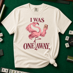 I Was One Away T-Shirt in Ivory - knotted tee flat lay on a green mahjong mat with tiles, printed with a pink flamingo standing on one leg and holding a mahjong tile, with the words I WAS ONE AWAY.