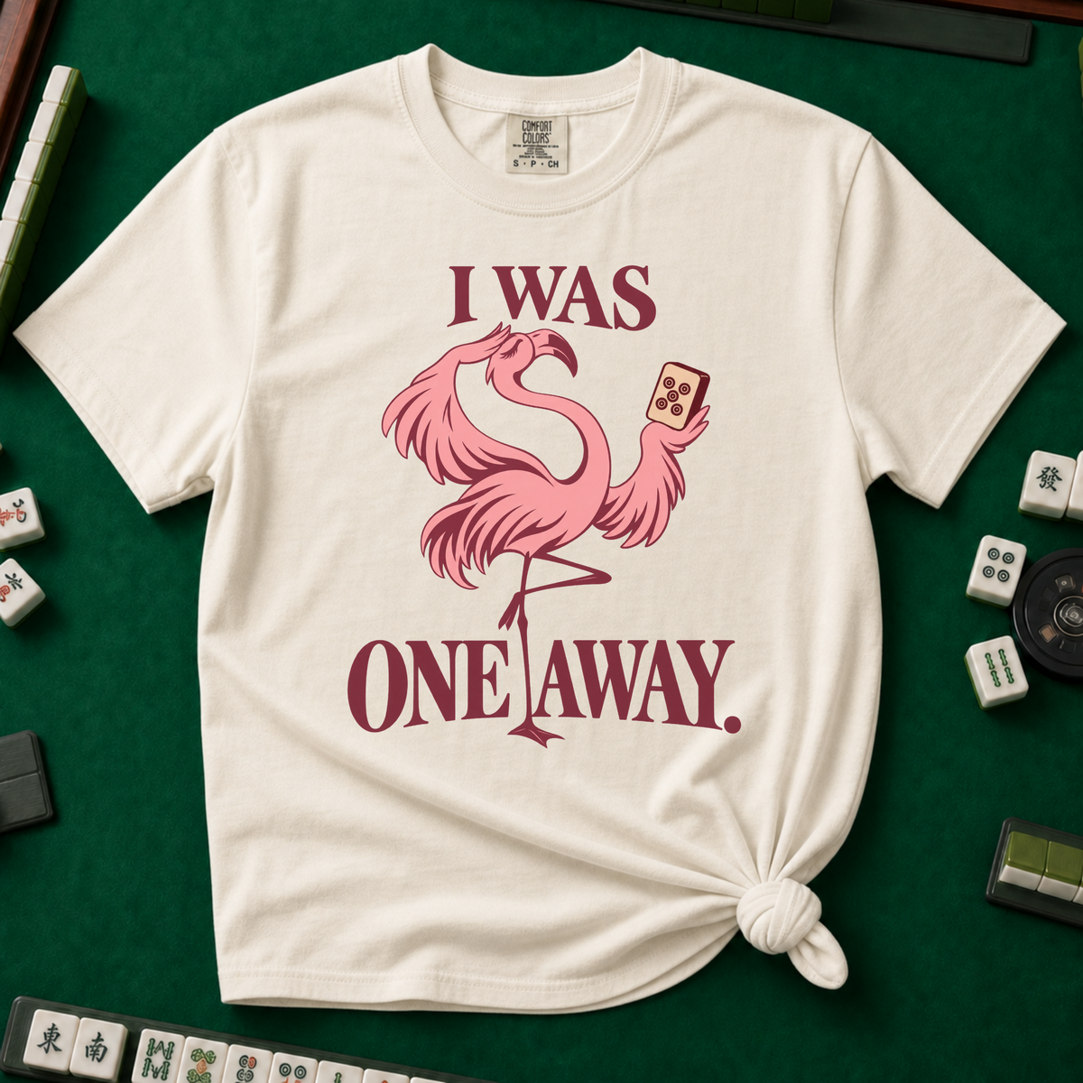I Was One Away T-Shirt in Ivory - knotted tee flat lay on a green mahjong mat with tiles, printed with a pink flamingo standing on one leg and holding a mahjong tile, with the words I WAS ONE AWAY.