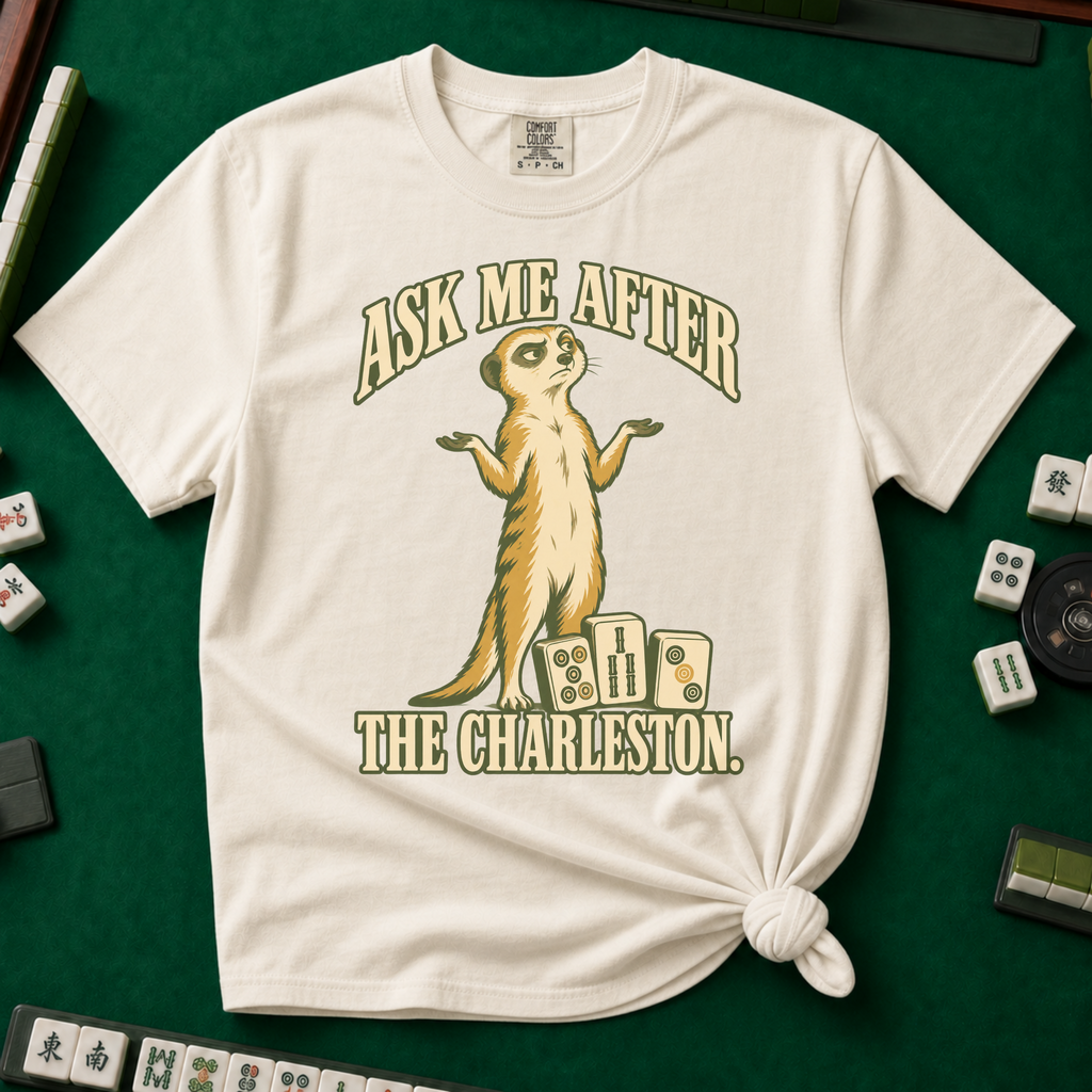 Ask Me After The Charleston T-Shirt in Ivory, knotted flat lay on a green mahjong mat - shrugging meerkat standing beside three mahjong tiles - "ASK ME AFTER THE CHARLESTON." in cream collegiate letters