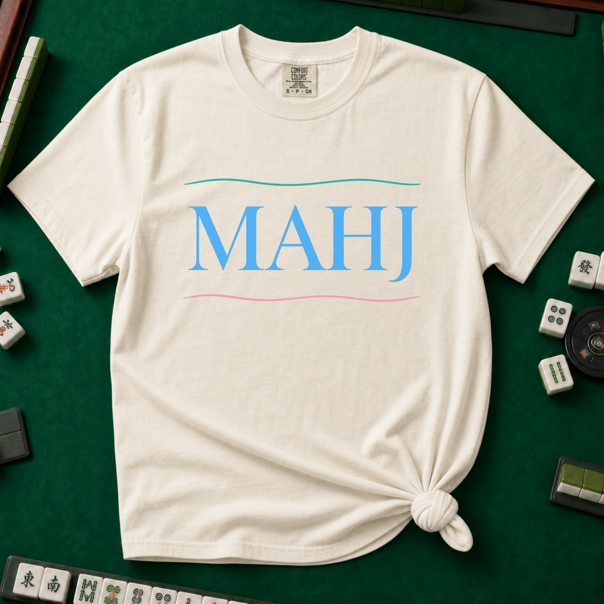 MAHJ Wave T-Shirt in Ivory, knotted flat lay on a green mahjong mat - "MAHJ" in sky-blue serif letters between a teal wave line above and a pink wave line below
