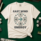 East Wind Energy T-Shirt in Ivory, knotted flat lay on a green mahjong mat - the four mahjong wind tiles arranged in a compass circle - North on top, West left, East right, South below - with a big teal arrow pointing East, between "EAST WIND" and "ENERGY" in charcoal block letters