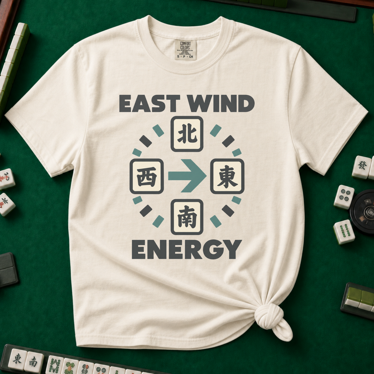 East Wind Energy T-Shirt in Ivory, knotted flat lay on a green mahjong mat - the four mahjong wind tiles arranged in a compass circle - North on top, West left, East right, South below - with a big teal arrow pointing East, between "EAST WIND" and "ENERGY" in charcoal block letters