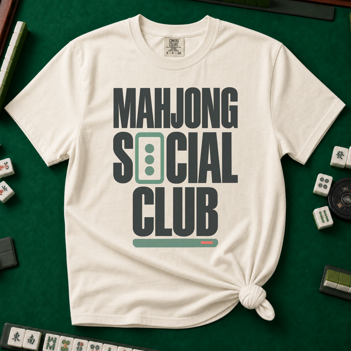 Mahjong Social Club Bold T-Shirt in Ivory, knotted flat lay on a green mahjong mat - bold charcoal block letters "MAHJONG SOCIAL CLUB" with a green three-dot tile as the O in SOCIAL and a green pusher bar underline
