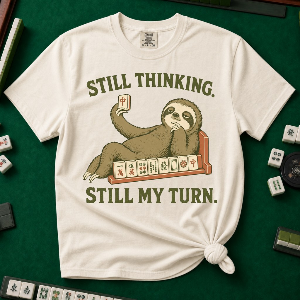 Still Thinking Still My Turn T-Shirt in Ivory, knotted flat lay on a green mahjong mat - a sloth draped along its mahjong tile rack, chin in hand, holding up a red dragon tile - "STILL THINKING. STILL MY TURN." in olive green vintage letters