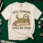 Still Thinking Still My Turn T-Shirt in Ivory, knotted flat lay on a green mahjong mat - a sloth draped along its mahjong tile rack, chin in hand, holding up a red dragon tile - "STILL THINKING. STILL MY TURN." in olive green vintage letters