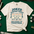 Joker Support Group Holding It Together T-Shirt in Ivory, knotted flat lay on a green mahjong mat - three retro cartoon joker tiles in sneakers, the middle one sweating nervously while the other two hold him up - "JOKER SUPPORT GROUP - HOLDING IT TOGETHER." in teal retro letters