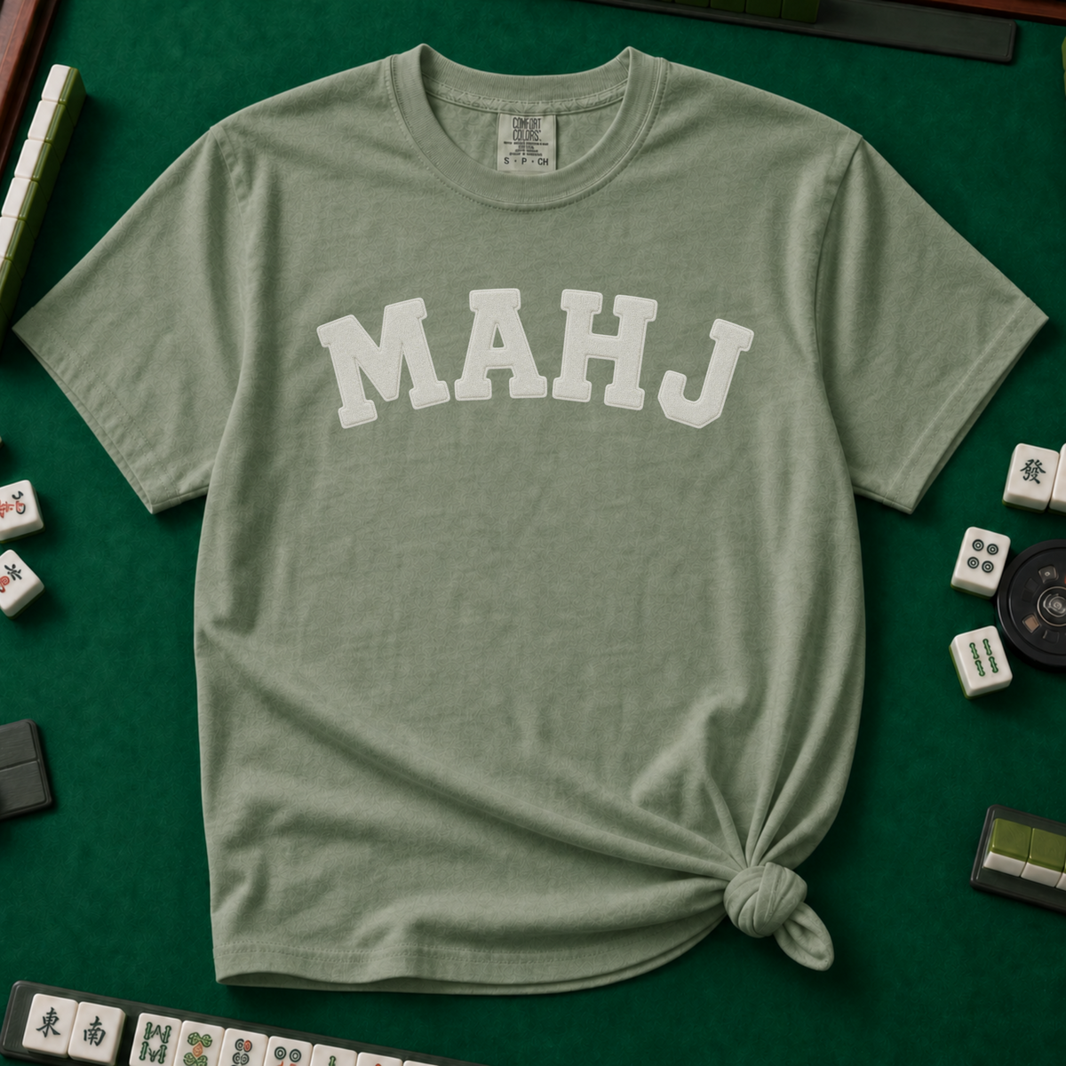 MAHJ Varsity T-Shirt in Moss, knotted flat lay on a green mahjong mat - "MAHJ" in big white varsity arch letters with a stitched letterman look