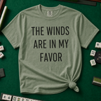 The Winds Are In My Favor T-Shirt in Moss, knotted flat lay on a green mahjong mat - "THE WINDS ARE IN MY FAVOR" in tall black letters