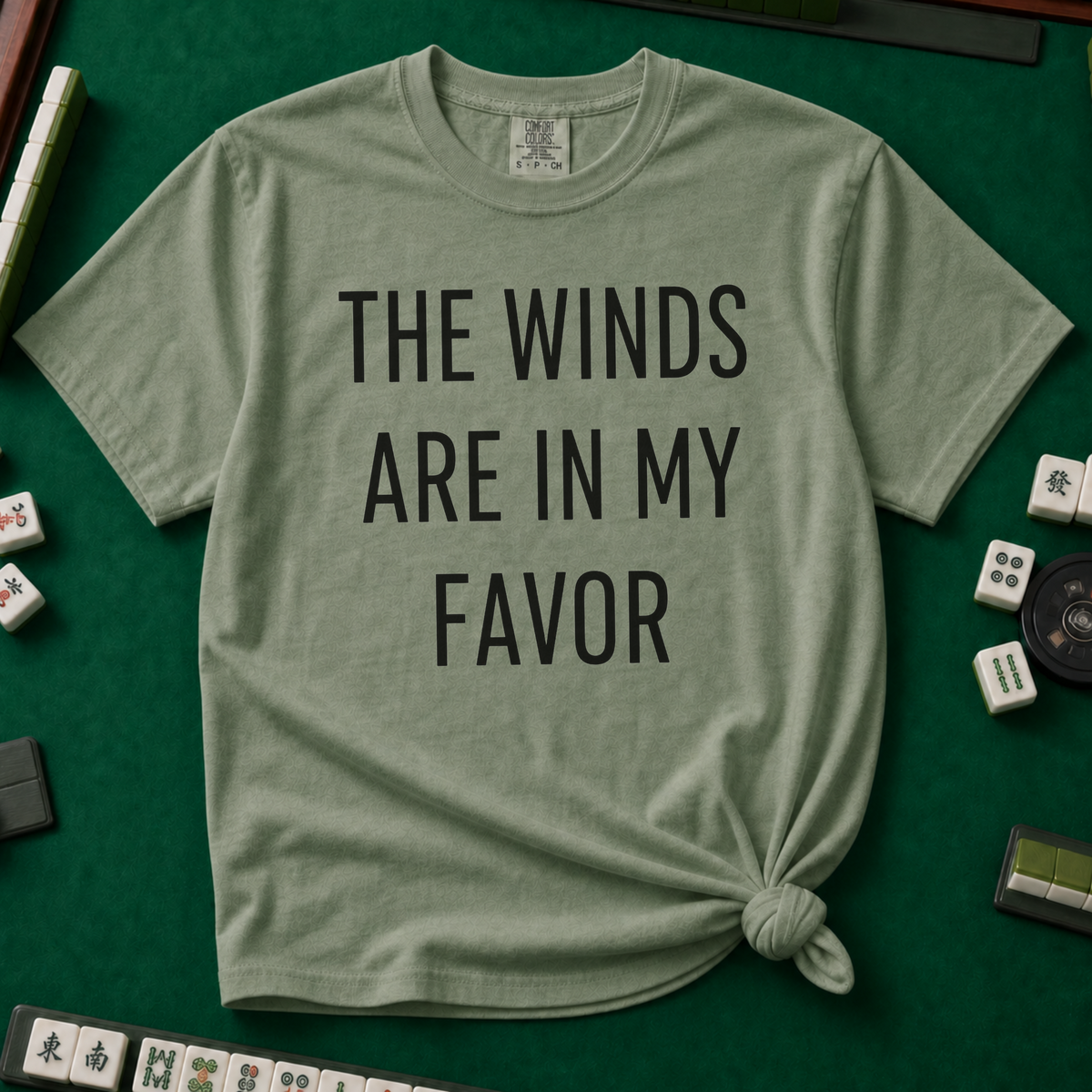 The Winds Are In My Favor T-Shirt in Moss, knotted flat lay on a green mahjong mat - "THE WINDS ARE IN MY FAVOR" in tall black letters