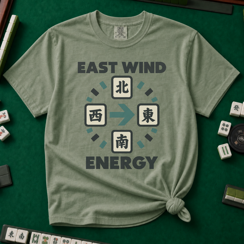 East Wind Energy T-Shirt in Moss, knotted flat lay on a green mahjong mat - the four mahjong wind tiles arranged in a compass circle - North on top, West left, East right, South below - with a big teal arrow pointing East, between "EAST WIND" and "ENERGY" in charcoal block letters