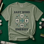 East Wind Energy T-Shirt in Moss, knotted flat lay on a green mahjong mat - the four mahjong wind tiles arranged in a compass circle - North on top, West left, East right, South below - with a big teal arrow pointing East, between "EAST WIND" and "ENERGY" in charcoal block letters