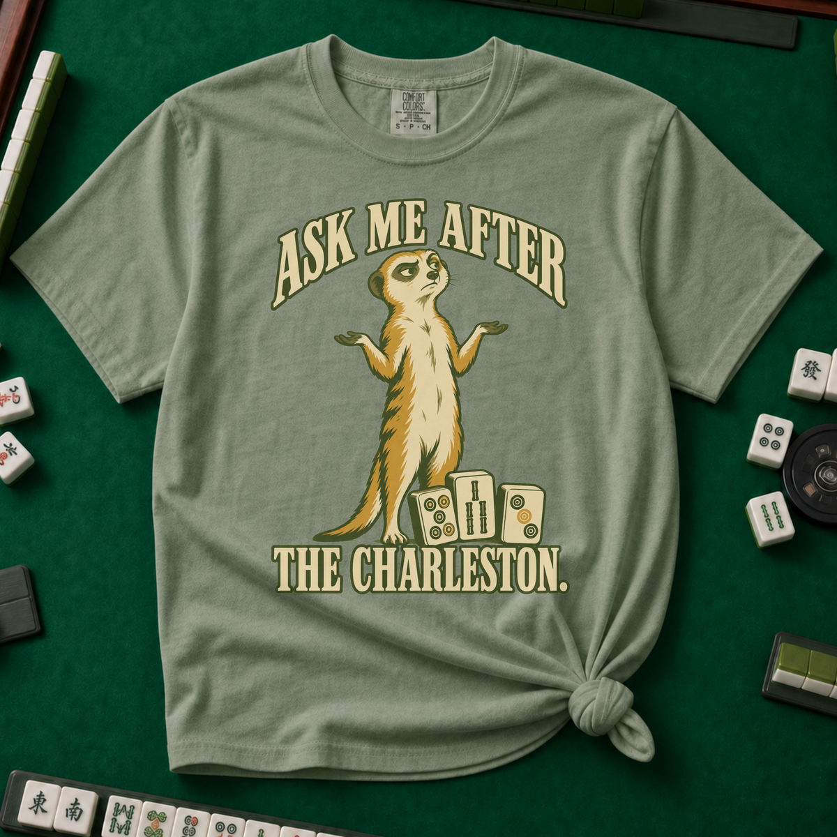Ask Me After The Charleston T-Shirt in Moss, knotted flat lay on a green mahjong mat - shrugging meerkat standing beside three mahjong tiles - "ASK ME AFTER THE CHARLESTON." in cream collegiate letters
