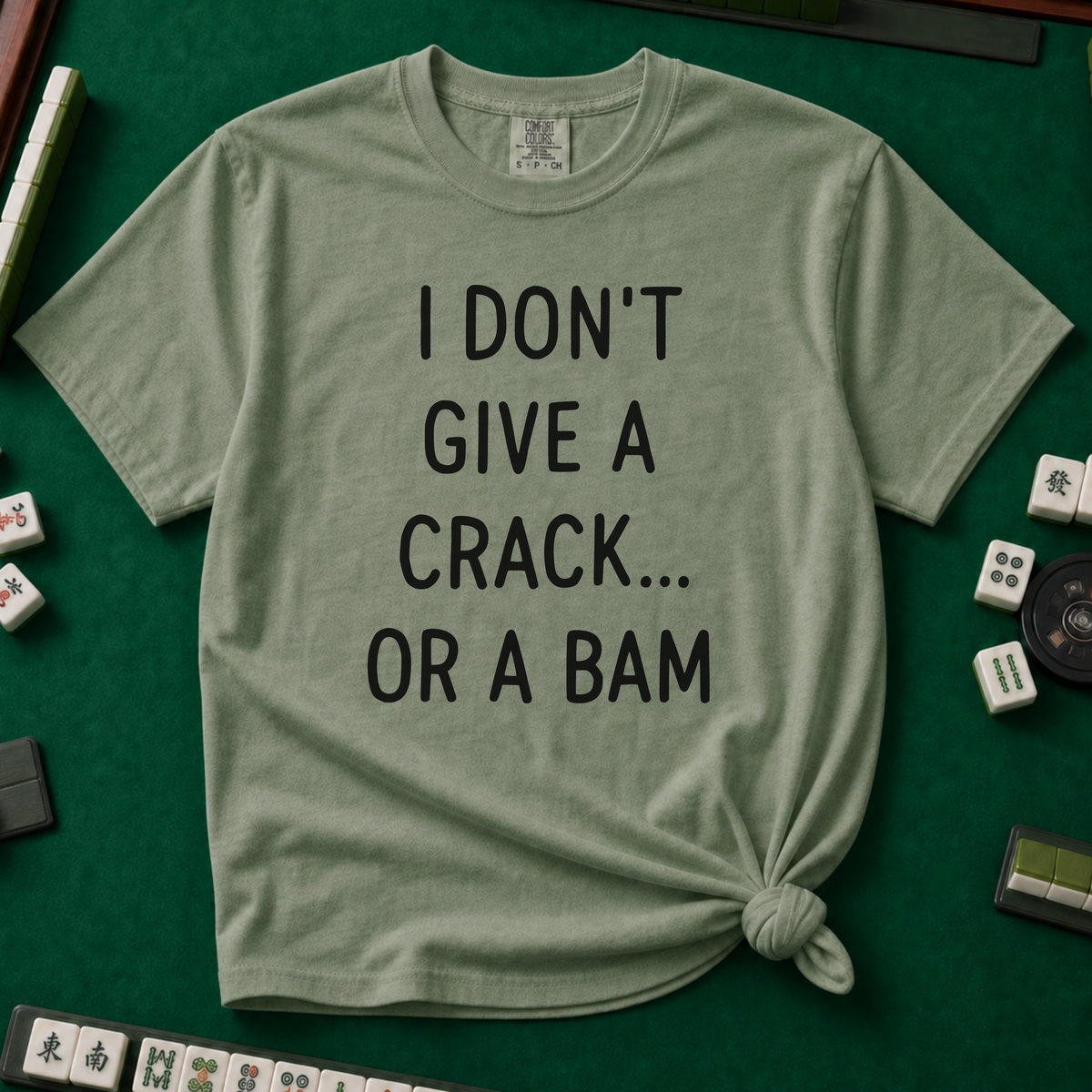 I Don't Give A Crack Or A Bam T-Shirt in Moss, knotted flat lay on a green mahjong mat - "I DON'T GIVE A CRACK... OR A BAM" in black handwritten-style letters