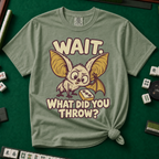 Wait What Did You Throw T-Shirt in Moss, knotted flat lay on a green mahjong mat - a wide-eyed fluffy bat with huge golden ears leaning in over a fallen dot tile - "WAIT. WHAT DID YOU THROW?" in cream bubble letters