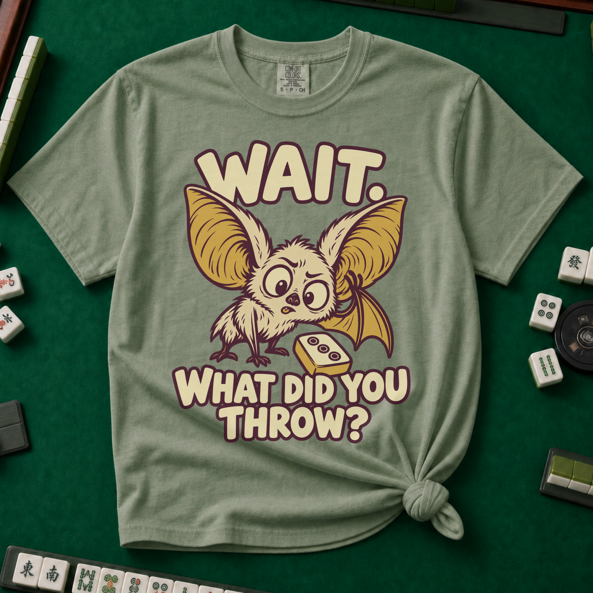 Wait What Did You Throw T-Shirt in Moss, knotted flat lay on a green mahjong mat - a wide-eyed fluffy bat with huge golden ears leaning in over a fallen dot tile - "WAIT. WHAT DID YOU THROW?" in cream bubble letters