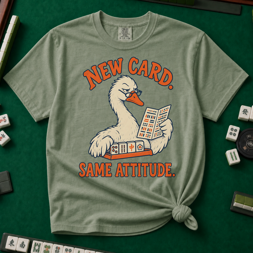 New Card T-Shirt in Moss - knotted tee flat lay on a green mahjong mat with tiles, printed with a white goose in glasses reading the new mahjong card over a rack of tiles, with the words NEW CARD. SAME ATTITUDE.