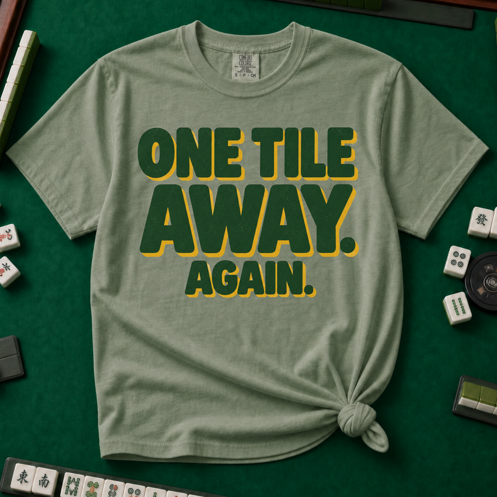 One Tile Away Again T-Shirt in Moss, knotted flat lay on a green mahjong mat - "ONE TILE AWAY. AGAIN." in bold deep-green block letters with a gold drop shadow