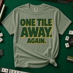 One Tile Away Again T-Shirt in Moss, knotted flat lay on a green mahjong mat - "ONE TILE AWAY. AGAIN." in bold deep-green block letters with a gold drop shadow