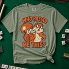 Who Passed Me This T-Shirt in Moss, knotted flat lay on a green mahjong mat - retro orange squirrel holding three mahjong tiles, pointing an accusing paw - "WHO PASSED ME THIS?" in bold orange letters