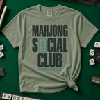 Mahjong Social Club Bold T-Shirt in Moss, knotted flat lay on a green mahjong mat - bold charcoal block letters "MAHJONG SOCIAL CLUB" with a green three-dot tile as the O in SOCIAL and a green pusher bar underline