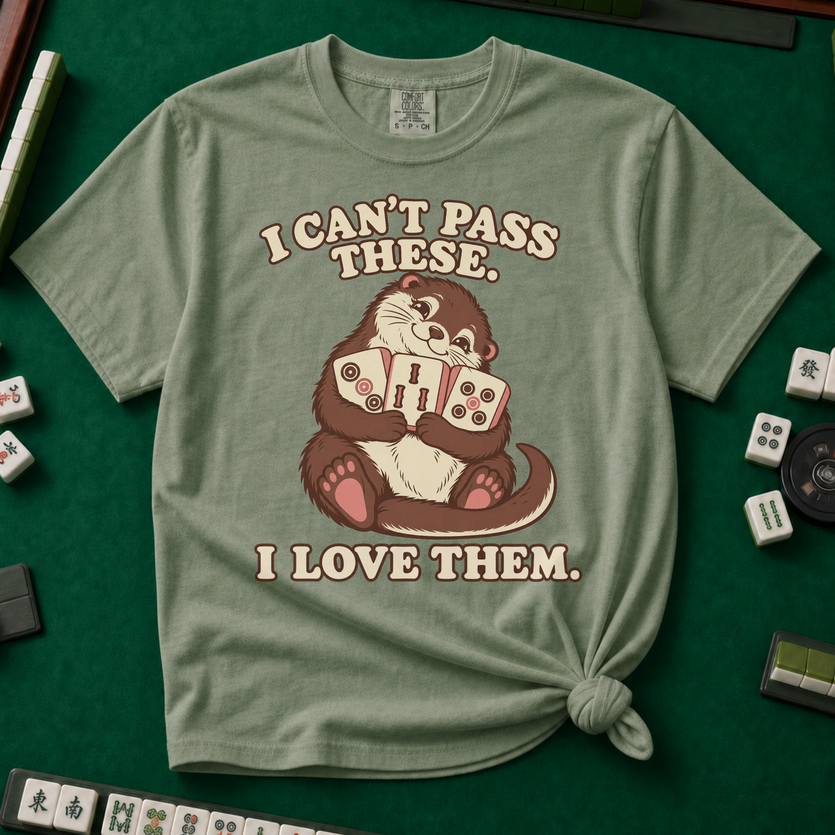 I Can't Pass These I Love Them T-Shirt in Moss, knotted flat lay on a green mahjong mat - lovestruck otter hugging three mahjong tiles to her chest - "I CAN'T PASS THESE. I LOVE THEM." in cream retro letters