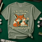 I'm Trying To Look Disappointed T-Shirt in Moss, knotted flat lay on a green mahjong mat - smug retro fox holding up a joker tile, hiding a grin behind one paw, tile rack in front - "I'M TRYING TO LOOK DISAPPOINTED." in cream collegiate letters