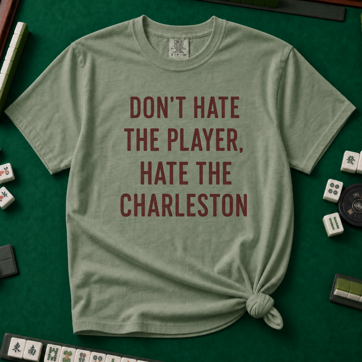 Don't Hate The Player Hate The Charleston T-Shirt in Moss, knotted flat lay on a green mahjong mat - "DON'T HATE THE PLAYER, HATE THE CHARLESTON" in deep burgundy block letters