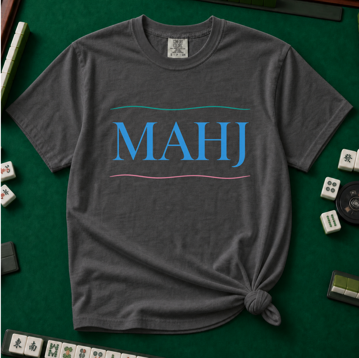 MAHJ Wave T-Shirt in Pepper, knotted flat lay on a green mahjong mat - "MAHJ" in sky-blue serif letters between a teal wave line above and a pink wave line below