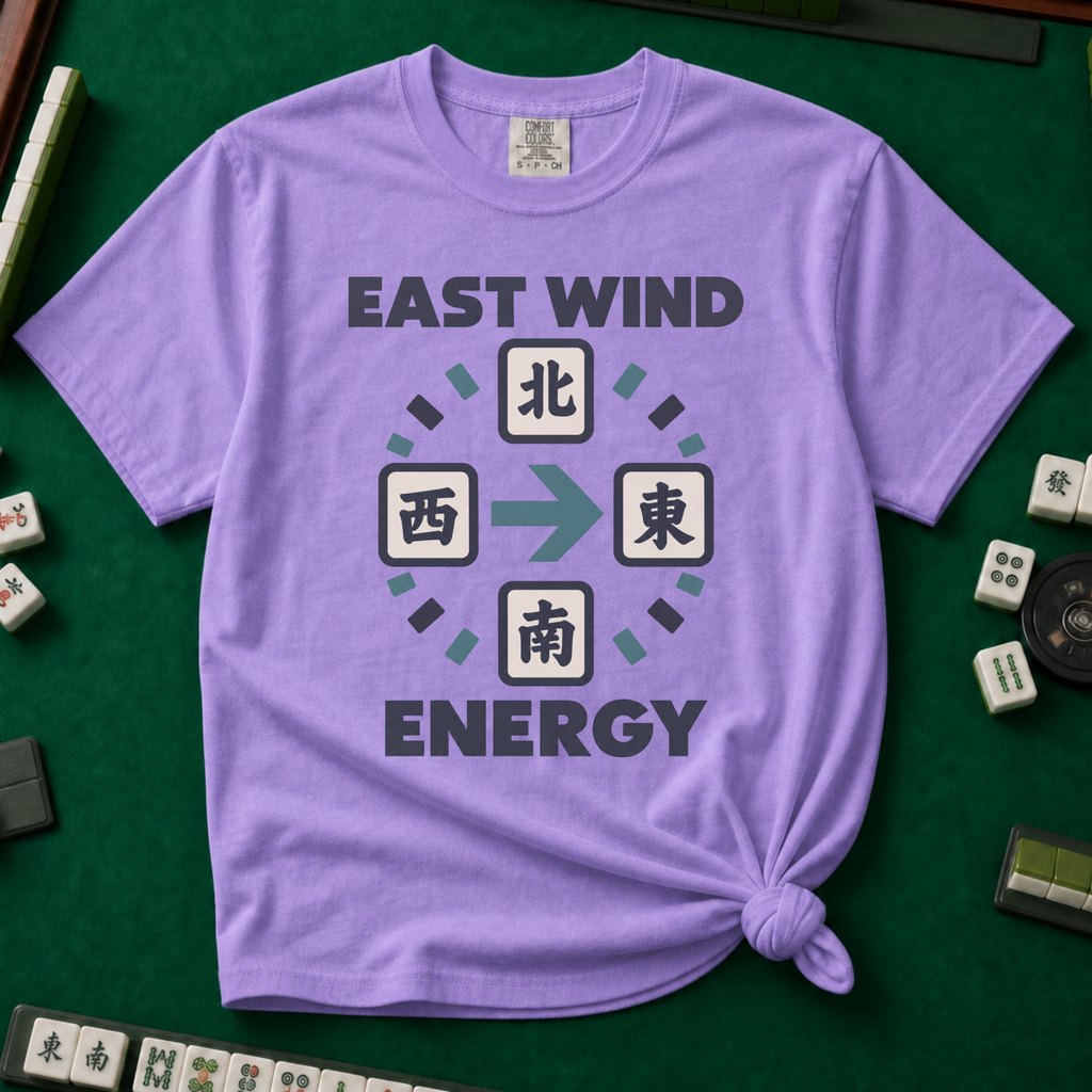 East Wind Energy T-Shirt in Violet, knotted flat lay on a green mahjong mat - the four mahjong wind tiles arranged in a compass circle - North on top, West left, East right, South below - with a big teal arrow pointing East, between "EAST WIND" and "ENERGY" in charcoal block letters