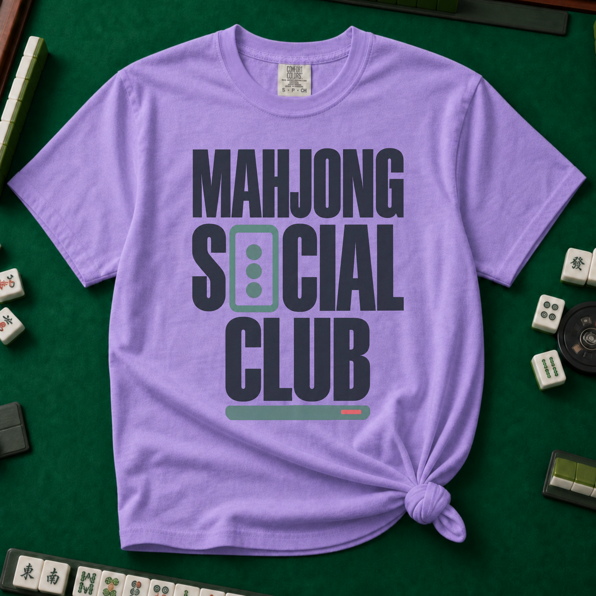 Mahjong Social Club Bold T-Shirt in Violet, knotted flat lay on a green mahjong mat - bold charcoal block letters "MAHJONG SOCIAL CLUB" with a green three-dot tile as the O in SOCIAL and a green pusher bar underline