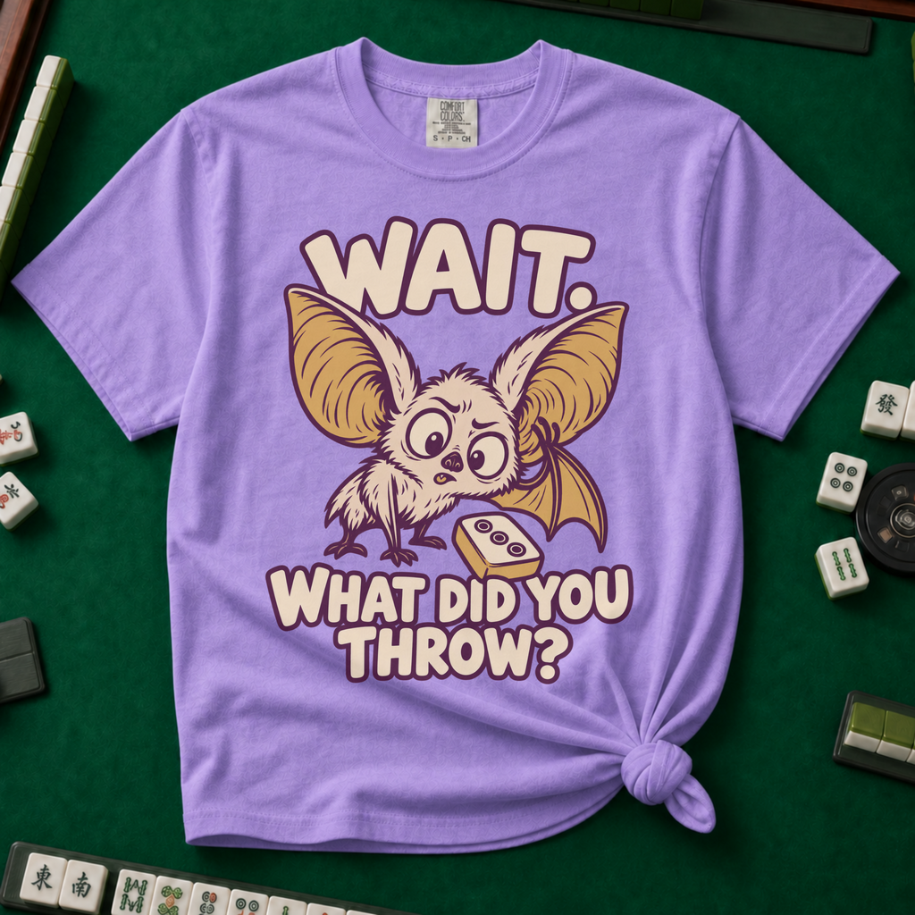 Wait What Did You Throw T-Shirt in Violet, knotted flat lay on a green mahjong mat - a wide-eyed fluffy bat with huge golden ears leaning in over a fallen dot tile - "WAIT. WHAT DID YOU THROW?" in cream bubble letters