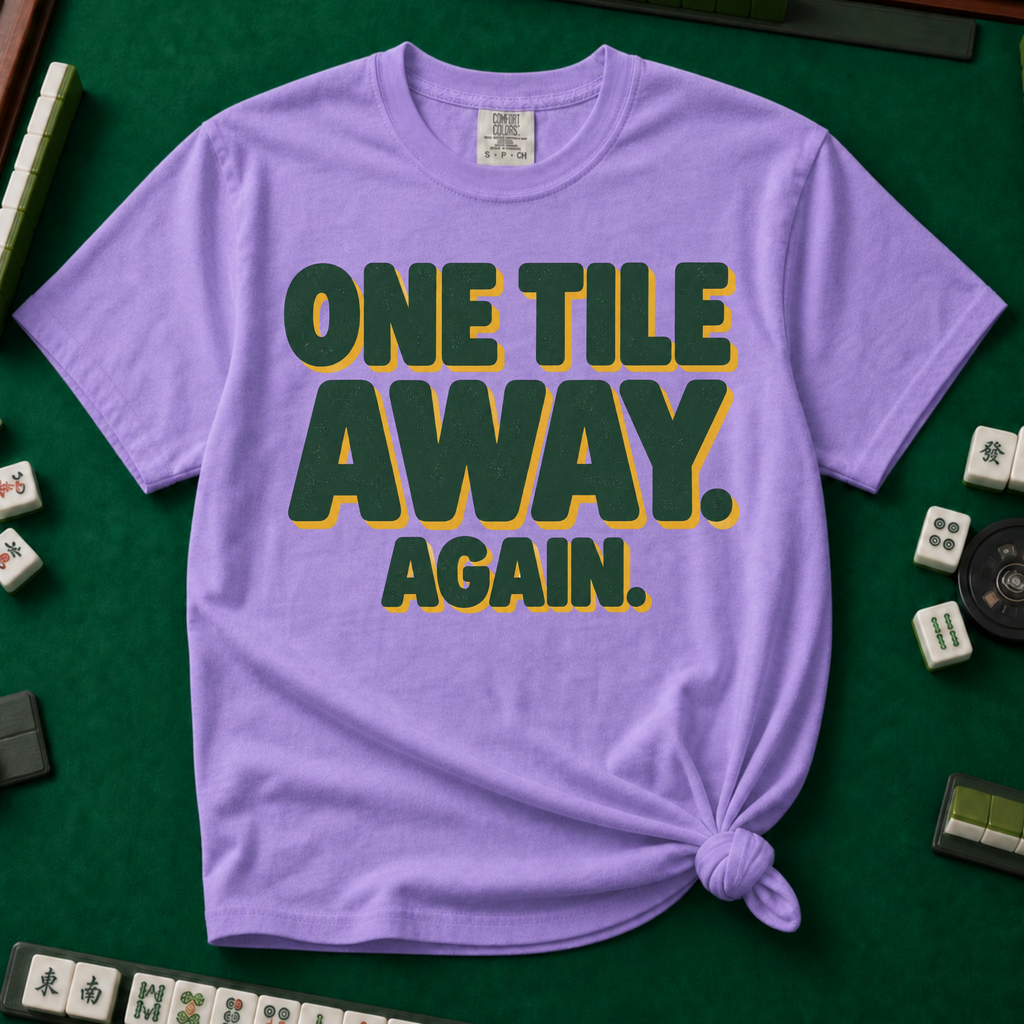 One Tile Away Again T-Shirt in Violet, knotted flat lay on a green mahjong mat - "ONE TILE AWAY. AGAIN." in bold deep-green block letters with a gold drop shadow