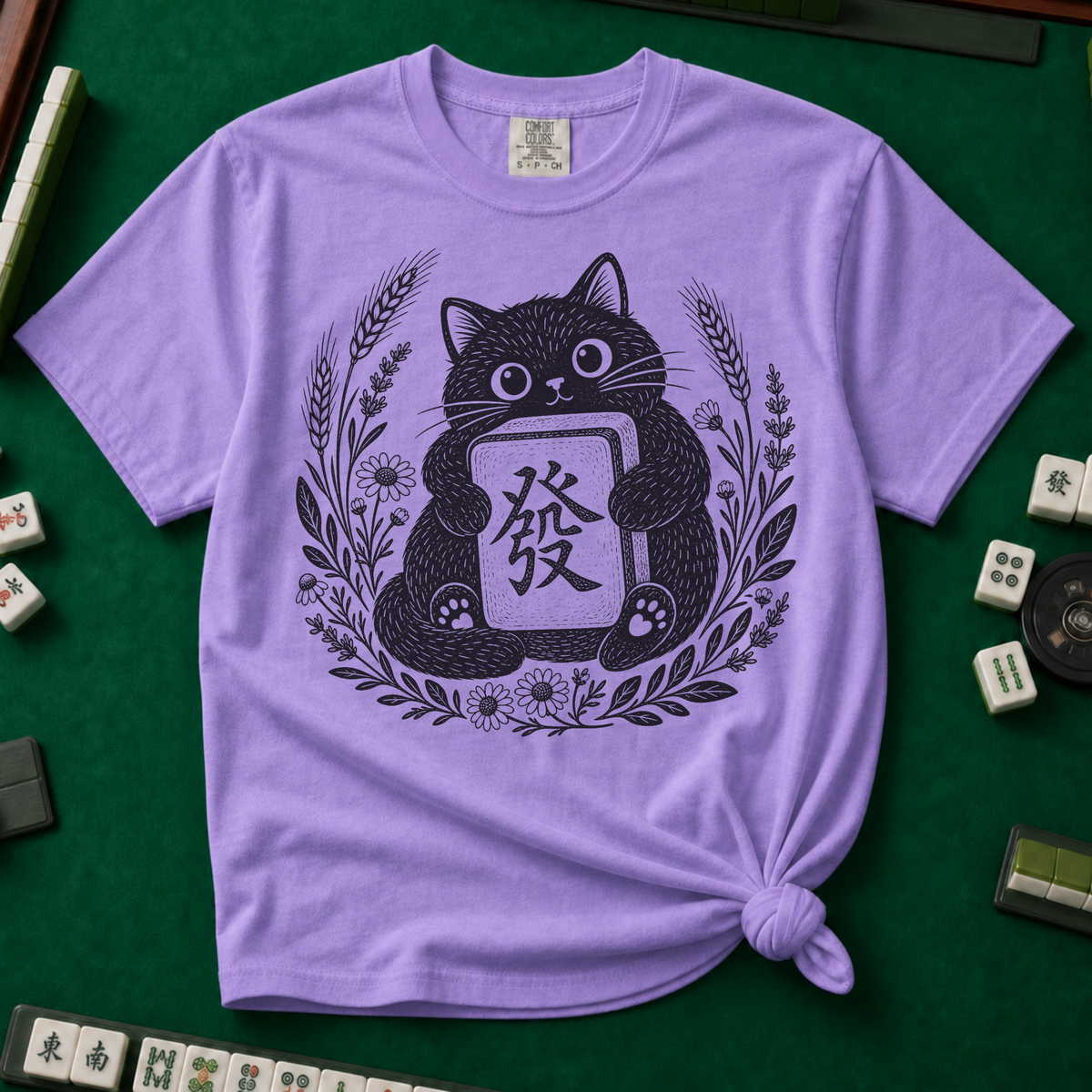 Linocut Tile Cat T-Shirt in Violet, knotted flat lay on a green mahjong mat - a linocut-style black cat hugging a Green Dragon mahjong tile inside a wreath of wheat, daisies and lavender