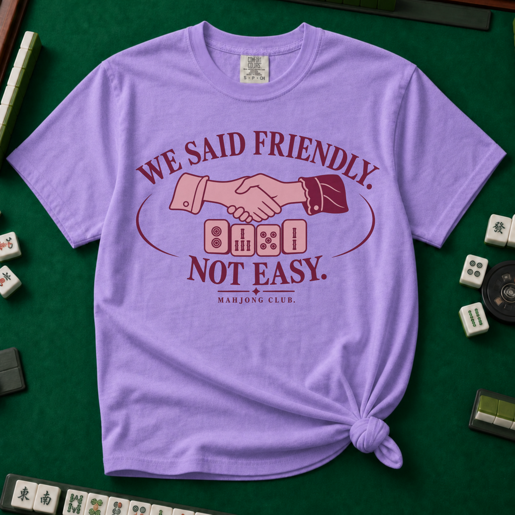We Said Friendly Not Easy T-Shirt in Violet, knotted flat lay on a green mahjong mat - a burgundy and pink club badge with a handshake over four mahjong tiles and the words "WE SAID FRIENDLY. NOT EASY. MAHJONG CLUB."