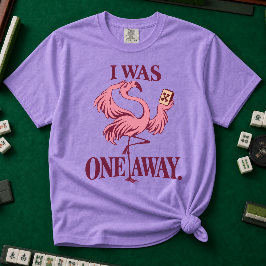 I Was One Away T-Shirt in Violet - knotted tee flat lay on a green mahjong mat with tiles, printed with a pink flamingo standing on one leg and holding a mahjong tile, with the words I WAS ONE AWAY.