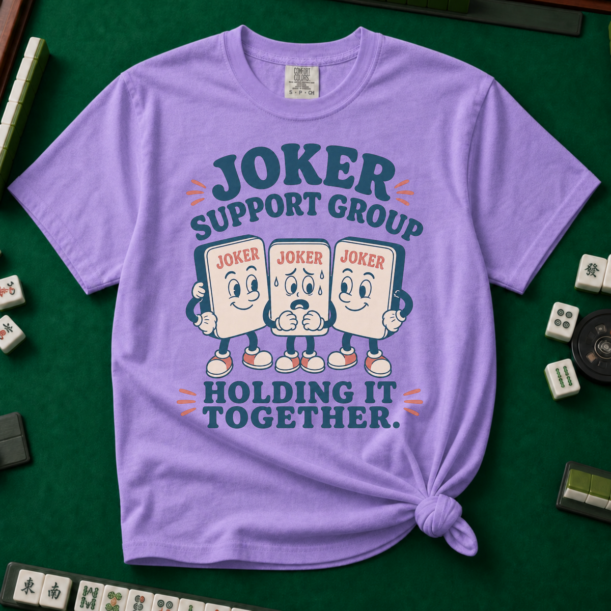 Joker Support Group Holding It Together T-Shirt in Violet, knotted flat lay on a green mahjong mat - three retro cartoon joker tiles in sneakers, the middle one sweating nervously while the other two hold him up - "JOKER SUPPORT GROUP - HOLDING IT TOGETHER." in teal retro letters