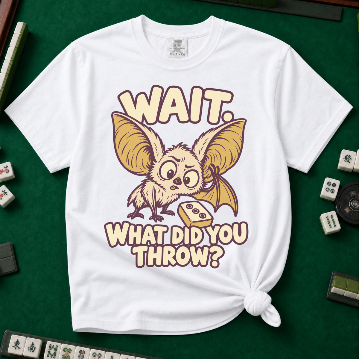 Wait What Did You Throw T-Shirt in White, knotted flat lay on a green mahjong mat - a wide-eyed fluffy bat with huge golden ears leaning in over a fallen dot tile - "WAIT. WHAT DID YOU THROW?" in cream bubble letters
