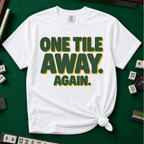 One Tile Away Again T-Shirt in White, knotted flat lay on a green mahjong mat - "ONE TILE AWAY. AGAIN." in bold deep-green block letters with a gold drop shadow