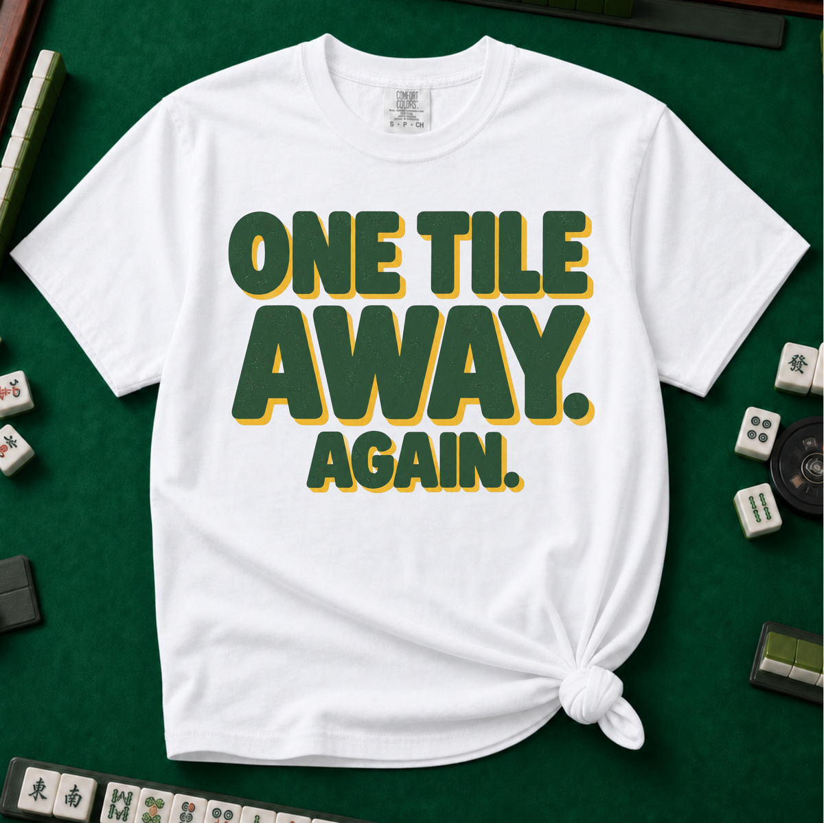 One Tile Away Again T-Shirt in White, knotted flat lay on a green mahjong mat - "ONE TILE AWAY. AGAIN." in bold deep-green block letters with a gold drop shadow