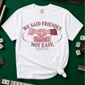 We Said Friendly Not Easy T-Shirt in White, knotted flat lay on a green mahjong mat - a burgundy and pink club badge with a handshake over four mahjong tiles and the words "WE SAID FRIENDLY. NOT EASY. MAHJONG CLUB."