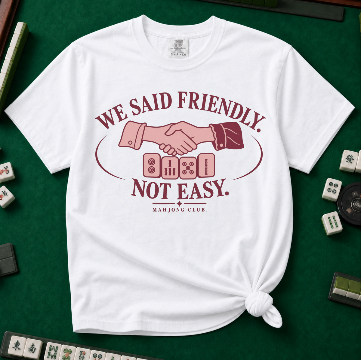 We Said Friendly Not Easy T-Shirt in White, knotted flat lay on a green mahjong mat - a burgundy and pink club badge with a handshake over four mahjong tiles and the words "WE SAID FRIENDLY. NOT EASY. MAHJONG CLUB."
