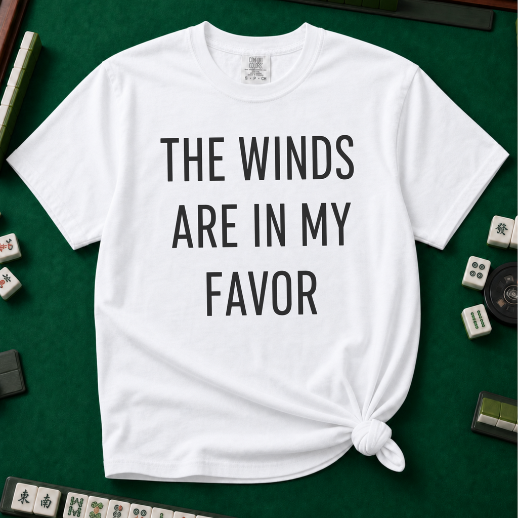 The Winds Are In My Favor T-Shirt in White, knotted flat lay on a green mahjong mat - "THE WINDS ARE IN MY FAVOR" in tall black letters