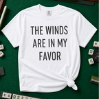 The Winds Are In My Favor T-Shirt in White, knotted flat lay on a green mahjong mat - "THE WINDS ARE IN MY FAVOR" in tall black letters