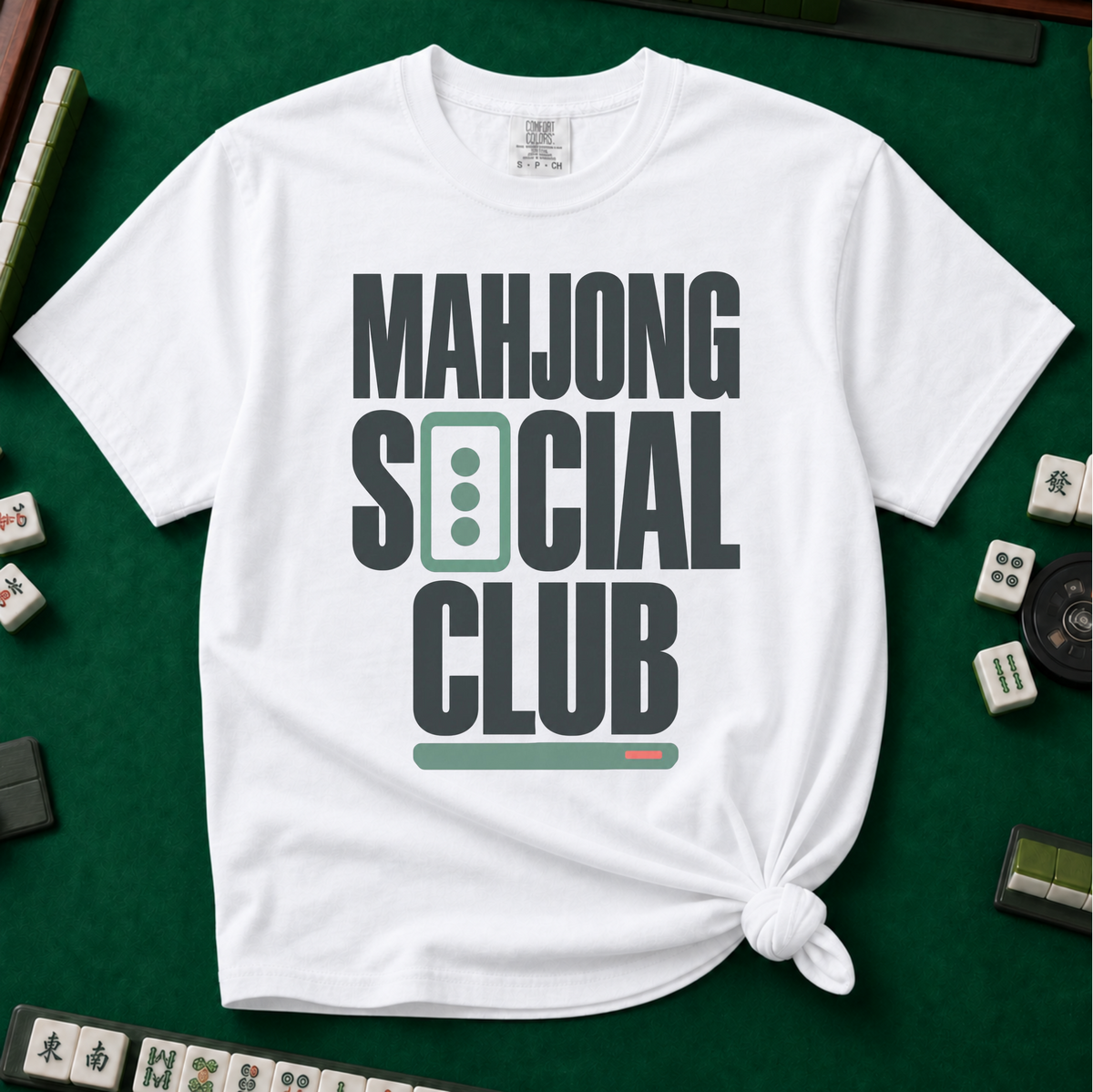 Mahjong Social Club Bold T-Shirt in White, knotted flat lay on a green mahjong mat - bold charcoal block letters "MAHJONG SOCIAL CLUB" with a green three-dot tile as the O in SOCIAL and a green pusher bar underline