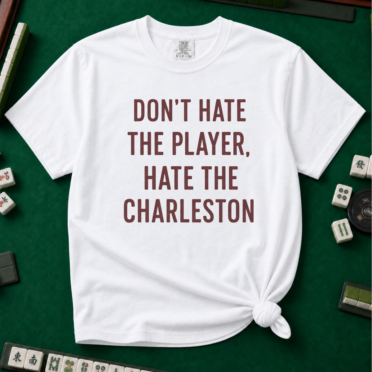 Don't Hate The Player Hate The Charleston T-Shirt in White, knotted flat lay on a green mahjong mat - "DON'T HATE THE PLAYER, HATE THE CHARLESTON" in deep burgundy block letters